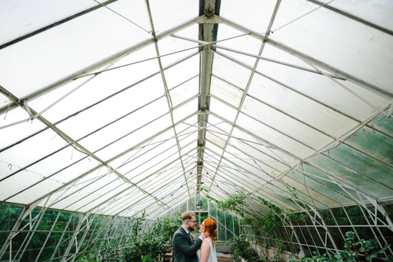 Happy just married couple lean in for a kiss inside a greenhouse by Jake Timms Photography - How To Look Your Best In Wedding Photos
