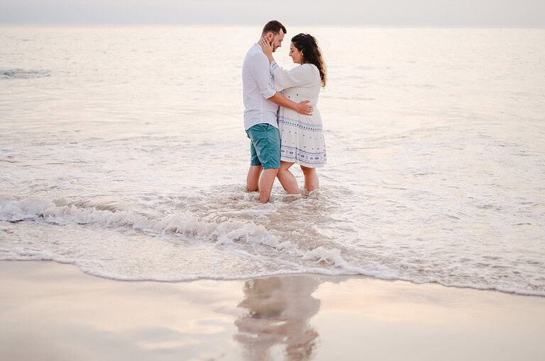 The waves wrap around them as the happy couple stand in the water as the sand mirrors them at Godrevy, Cornwall
