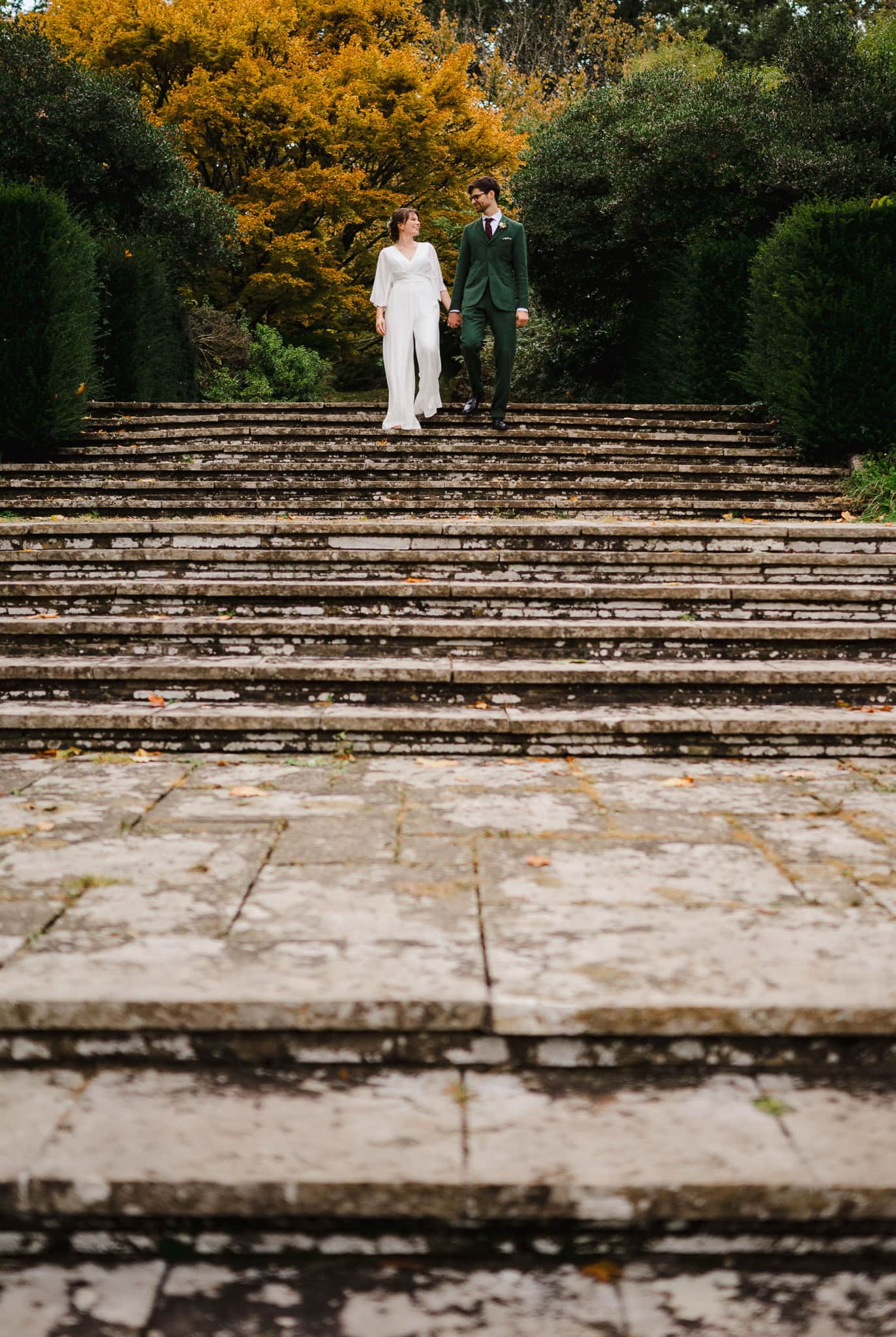 Happy couple walk down the steps at Darlington Hall surrounded by beautiful autumnal trees by Devon Wedding Photographer, Jake Timms Photography
